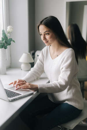 Serious focused young woman freelancer is typing on laptop sitting next to a window in the kitchen. Concept of everyday life.の写真素材