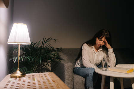 Young woman is thinking in the dark room. Girl sitting on the bed. Theres a desk lamp on the table.の写真素材