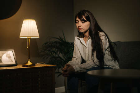 Ttired young woman sitting near a desk lamp in a dark room. Girls sitting there thinking about it.の写真素材