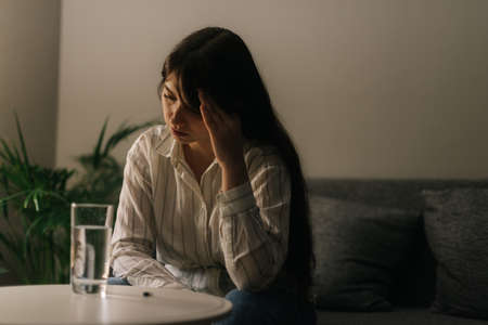 Sick tired beautiful woman sitting in front of a table with a tablet and a glass of water in a dark...