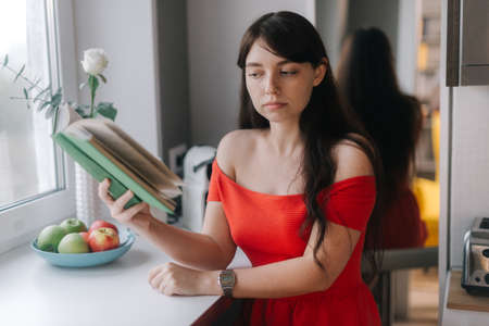Smiling young woman in red dress is reading a book by the window. Pretty girl in provocative bright clothes.の写真素材