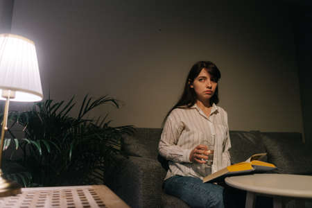 Beautiful female is holding a glass of water in hand in a dark room. On the table is a book. Woman looks away.の写真素材
