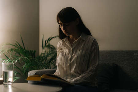 Tired young woman is reading a book in a dark room. Cute sick girl in casual clothes. The depressive look on a womans face.の写真素材