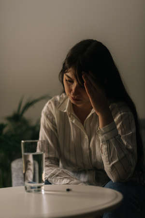 Unhealthy tired young woman sitting in front of a table with a tablet and a glass of water in a dark room.の写真素材