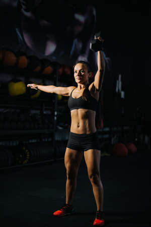 Fitness young woman with perfect muscular body in black sportswear is lifting kettlebell overhead during weight training workout. Concept of healthy lifestyle and workouts in a modern dark gym.の写真素材