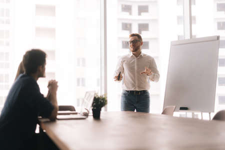 Young casual businessman presenting new project to partners on whiteboard in modern office meeting...