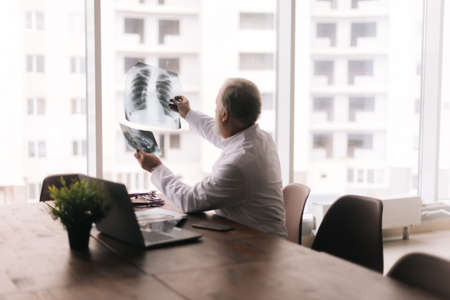 Adult male doctor examining x-ray and MRI scan at a table against large window in modern office room at hospital. Medical concept of the doctor's work at the workplace. Concept of medical work.の写真素材
