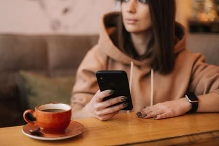 Close-up portrait of young attractive woman typing message in phone at cozy coffee shop. Pretty girl in casual clothes thinks while typing on the phone at cafe. Concept of leisure activity.の写真素材