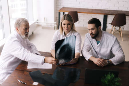 Group of three adult and young doctors discuss chest X-rays at desk against large window in conference room. Concept of team of doctors working together at the clinic. Concept of team medical work.の写真素材