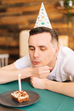 Close-up of face of man blowing out one candle on little cake. Smoke hovers beautifully in the air. Concept of celebrating alone.の写真素材