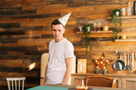 Upset young man in festive cup stands in cozy kitchen and looks at the cake on the table with sad eyes. Concept of celebrating alone.の写真素材