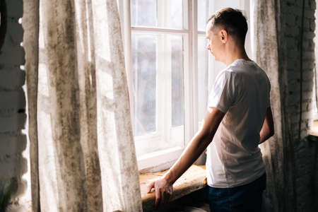 Thoughtful European young man stands by the window on sunny day, enjoying beautiful day.の写真素材
