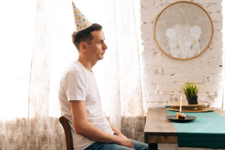 Upset young man sitting at the birthday cake and looking at away. Concept of solitude in quarantine...