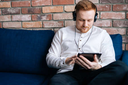 Caucasian young man in headphones watching movie on tablet computer while sitting on the couch at home. Concept of home relaxation.の写真素材