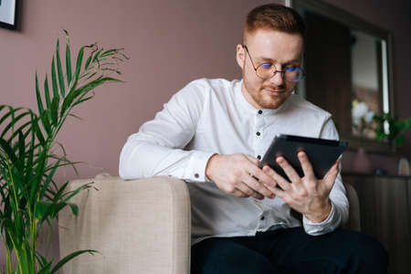 Stylish young man in fashion glasses sitting in soft armchair and using digital tablet at homeの写真素材