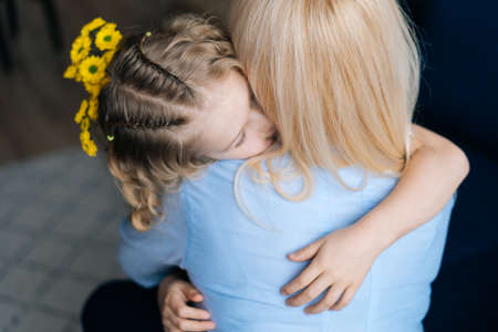 Blonde woman and her curly little daughter are hugging at the home in cozy living room. Loving...