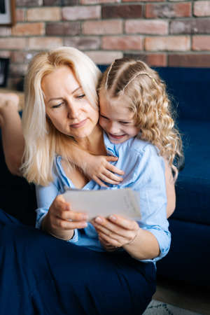 Blonde woman in casual clothes and her little curly daughter are looking through photos while sitting on couch at home in cozy living room. Concept of family holiday and togetherness.の写真素材