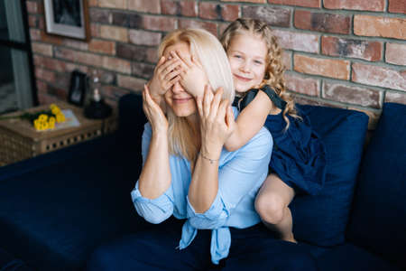 Charming curly daughter closes eyes to her mom while sitting on couch at home in cozy living room. Concept of family holiday and togetherness.の写真素材