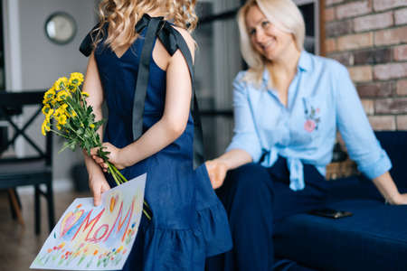Rear view of adorable daughter hiding behind back surprise for her mother. Girl gives her mother...