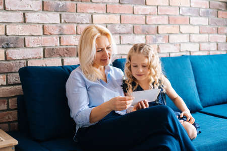 Beautiful blonde woman in casual clothes and her little curly daughter are looking through photos...