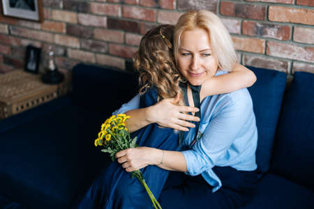 Beautiful blonde young woman and her curly charming little daughter are hugging at the home in cozy...
