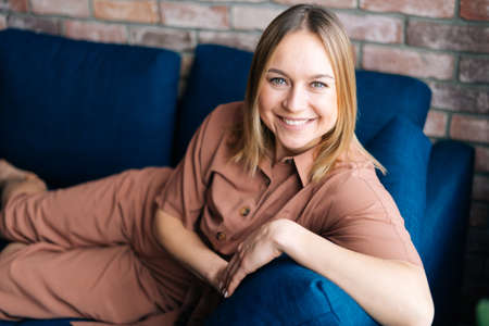 Young smiling blonde woman relaxing on the soft couch after long day, looking at the camera. Concept...