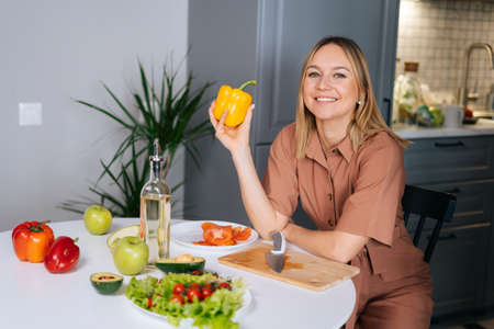 Beautiful young woman poses with yellow bell pepper at a table in kitchen with modern interior,...