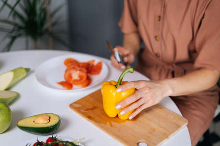 Close-up of hand of woman cutting fresh bell pepper using kitchen knife on wooden cutting board. Young woman cutting yellow bell pepper with a knife for vegetable salad. Concept of healthy eating.の写真素材