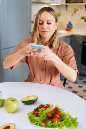 Young woman photographing fresh fruits and vegetables with smartphone in kitchen with modern...