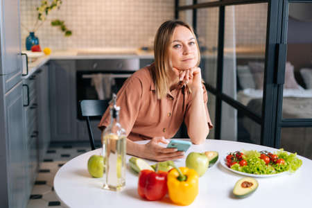 Pretty woman with fruits and mobile phone sitting at the table in kitchen with modern interior. Concept of healthy eating.の写真素材