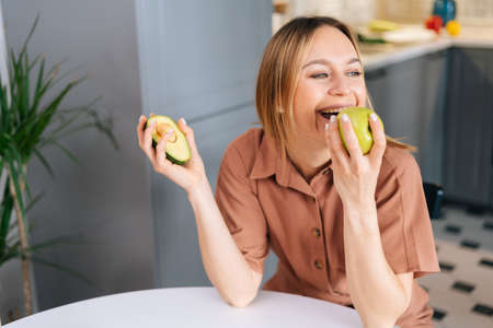 Portrait of young cheerful woman eating an apple at the table in the bright modern kitchen Concept of healthy eating.の写真素材