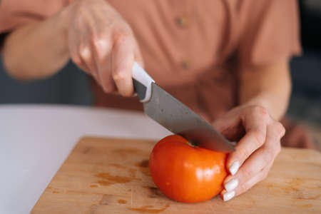 Close-up of hand of woman cutting fresh tomato using kitchen knife on wooden cutting board. Young...