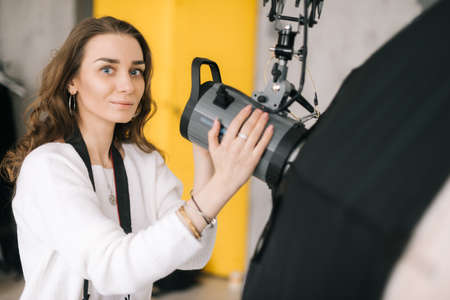 Portrait of young beautiful woman photographer prepares light equipment softbox for photo shoot in the studio, looking at camera. Concept of creative work in photo studio, backstage job.の写真素材