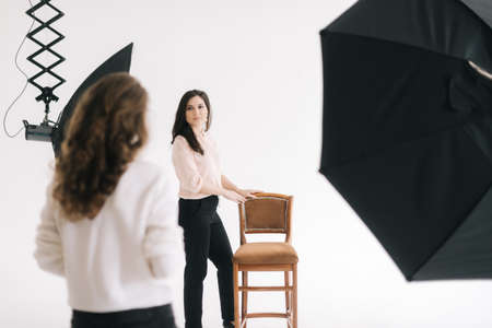 Attractive young female model posing with chair in photo studio for female photographer while she is...