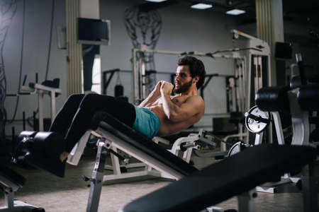 Sportive bearded young man with muscular wiry naked torso doing abdominal exercises on floor with...