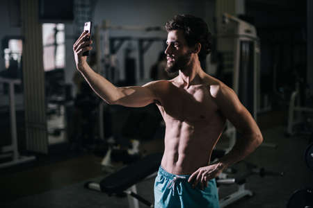 Handsome bearded young man with muscular wiry naked torso taking selfie photo on smartphone close-up looking camera during sport workout training in modern dark gym. Concept of healthy lifestyle.の写真素材