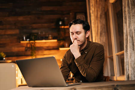 Thoughtful young man in casual clothing working on laptop while sitting at the table in cozy living...