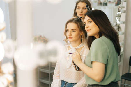 Young beautiful woman sitting in chair in front of mirror and gets a professional make-up in salon....