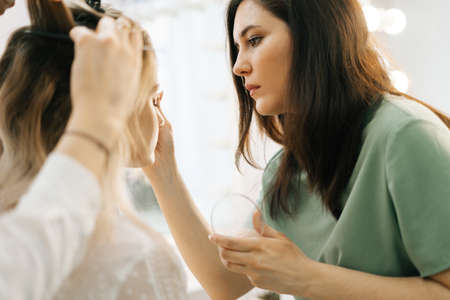 Young beautiful woman sitting in chair in front of mirror and gets a professional make-up in salon. Professional make-up artist works. Concept of backstage work.の写真素材