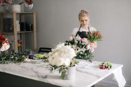 Professional young woman florist wearing apron making floral arrangement from fresh tulip at the table on white background. Concept of working with flowers, floral business.の写真素材