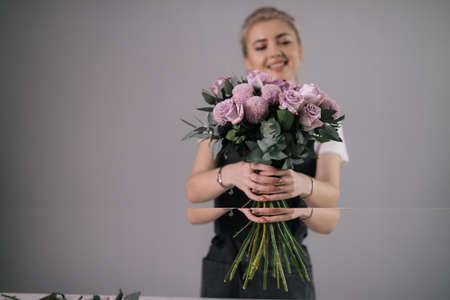 Smiling young woman florist wearing apron holding bouquet of fresh flowers in hands at the table on white background. Concept of working with flowers, floral business.の写真素材