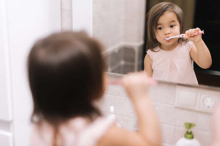 Cute beautiful little girl is brushing her teeth with tooth brush in front of the bathroom mirror, looking at the camera.の写真素材