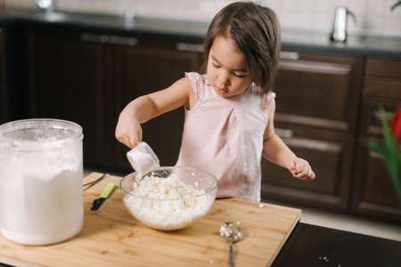 Beautiful little two-year-old girl wearing white dress is adding glass of flour to transparent bowl...