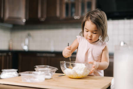 Cute beautiful little girl is kneading and mixing cottage cheese and eggs in transparent bowl for...