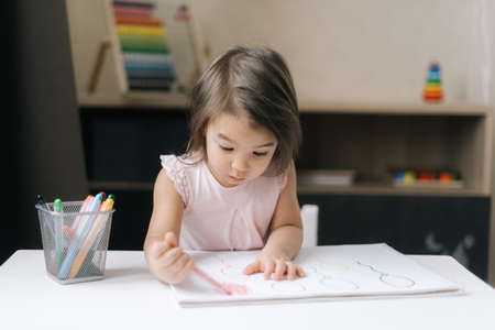 Adorable beautiful creative little girl is painting with felt-tip pens the figure of balloon on paper sitting at table in childrens room.の写真素材