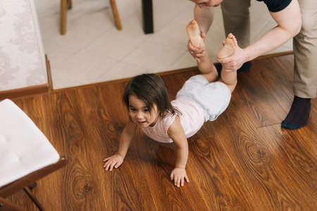 Happy father is playing with child holding his daughter legs who crawling on floor. Concept of carefree childhood.の写真素材