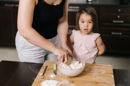 Moms kneading the dough for her little girl who makes cheesecakes at the table in kitchen with modern interior. .の写真素材