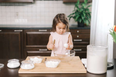 Little two-year-old girl is learning to bake cake, child standing breaking eggs into a bowl at...
