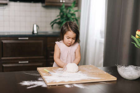 Cute beautiful little girl prepares dough for baking cookies at the table in kitchen with modern...