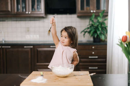 Asian beautiful little girl raises a spoon over her head while making cheesecakes dough at the table in kitchen with modern interior.の写真素材